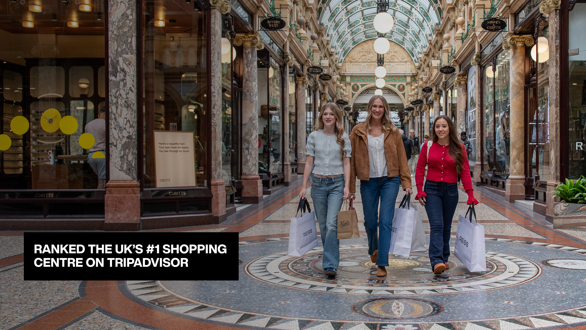 Shoppers walking through Victoria Leeds shopping arcade with luxury stores and glass ceiling.