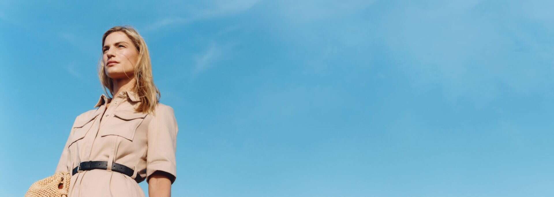 Elegant woman in a beige trench coat against a blue sky.