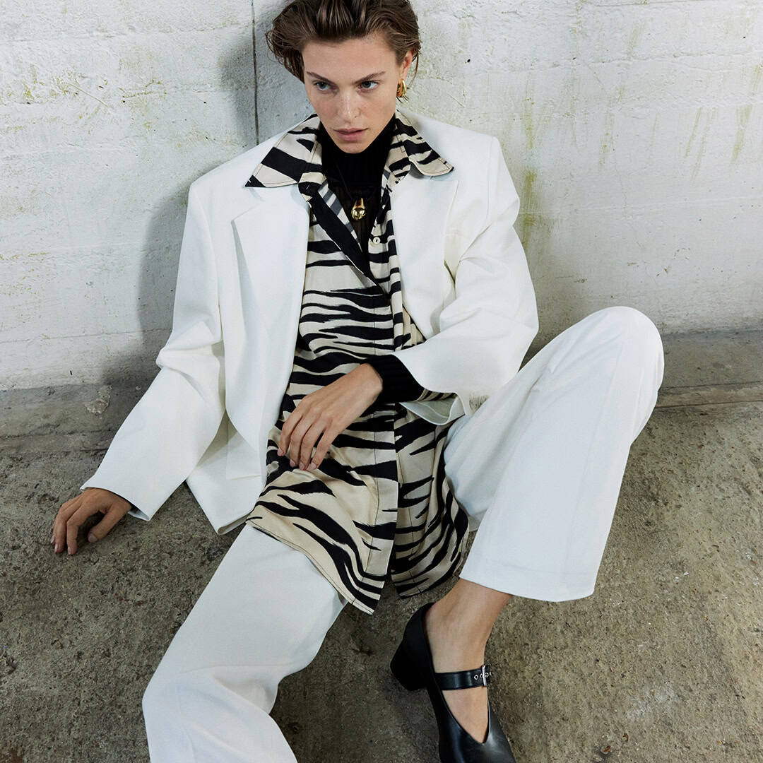 Woman in stylish black and white zebra print outfit with white blazer, sitting on concrete floor.