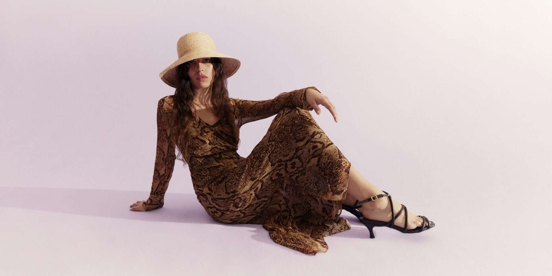 Elegant woman in a brown animal print dress with a wide-brim hat and black heels, sitting on the flo.
