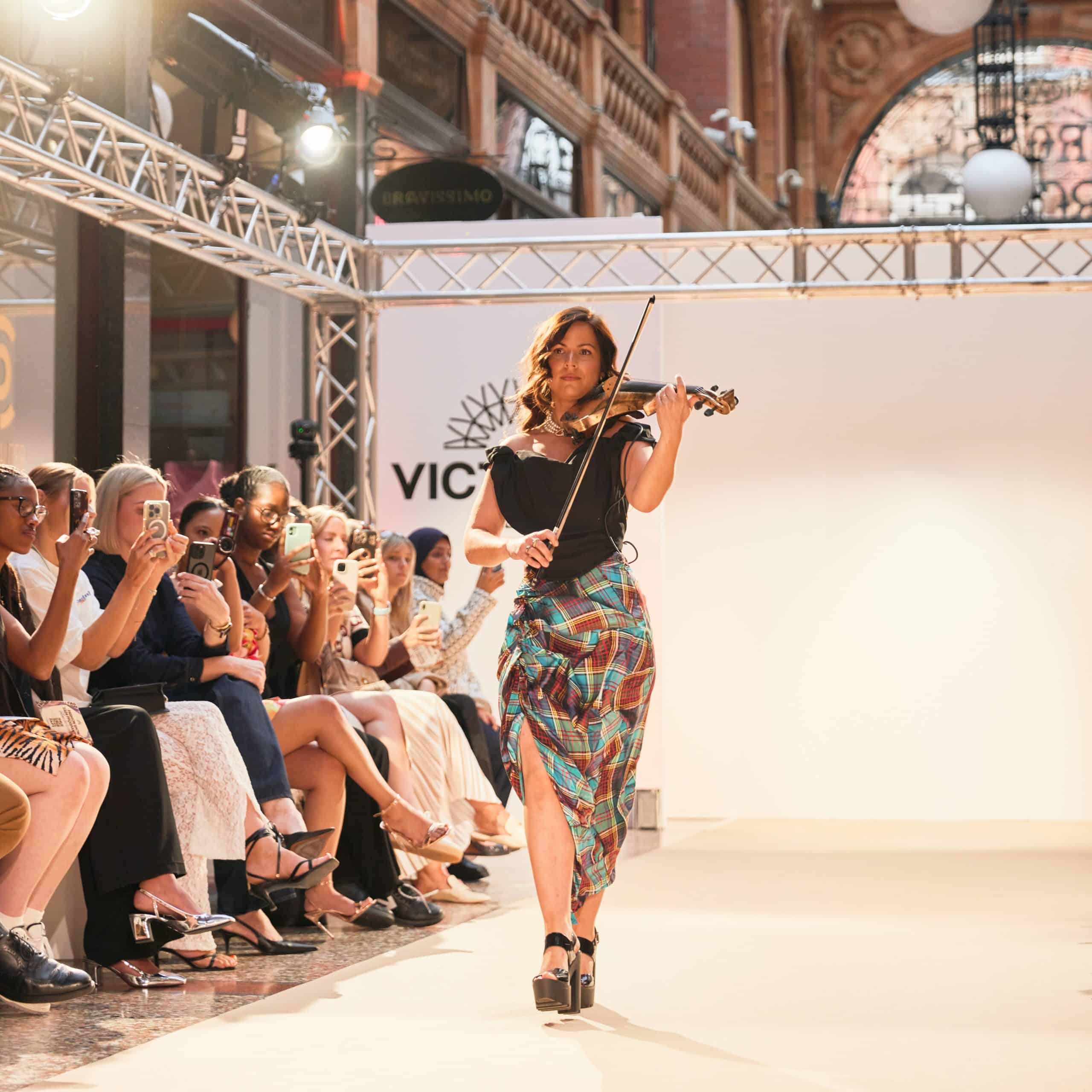Elegant woman walking on runway with violin at Victoria Leeds fashion event.