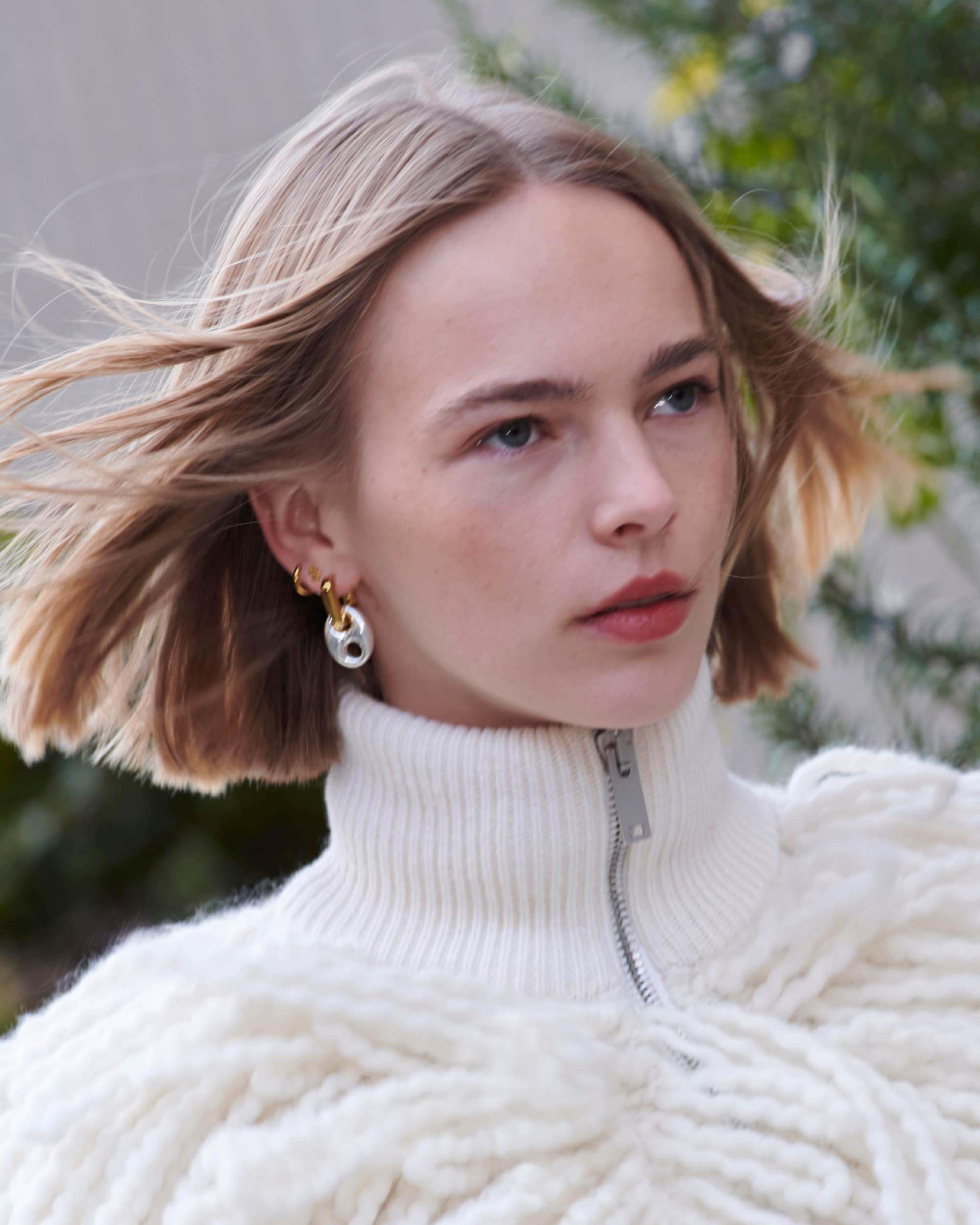 Elegant young woman with blonde hair and earrings, wearing a white zip-up sweater outdoors.