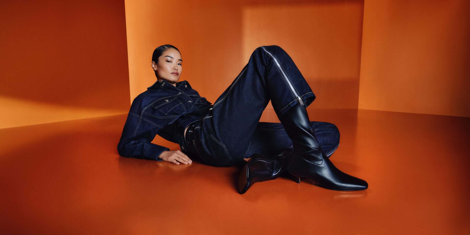Fashion model in dark denim outfit and black boots on orange background.